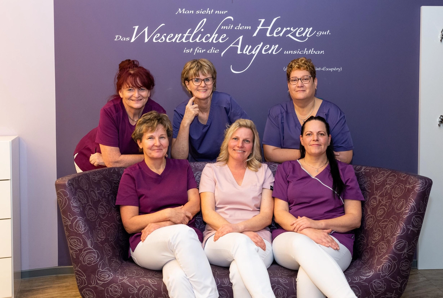 Frauenarztpraxis Dr. A. Dietrich - Gesundheitsdienste für Frauen - Eine Gruppe von sechs Frauen in lila und rosa OP-Kleidung posiert vor einer lila Wand mit deutscher Schrift. Drei Frauen stehen im Hintergrund, während drei auf einem gemusterten lila Sofa im Vordergrund sitzen.